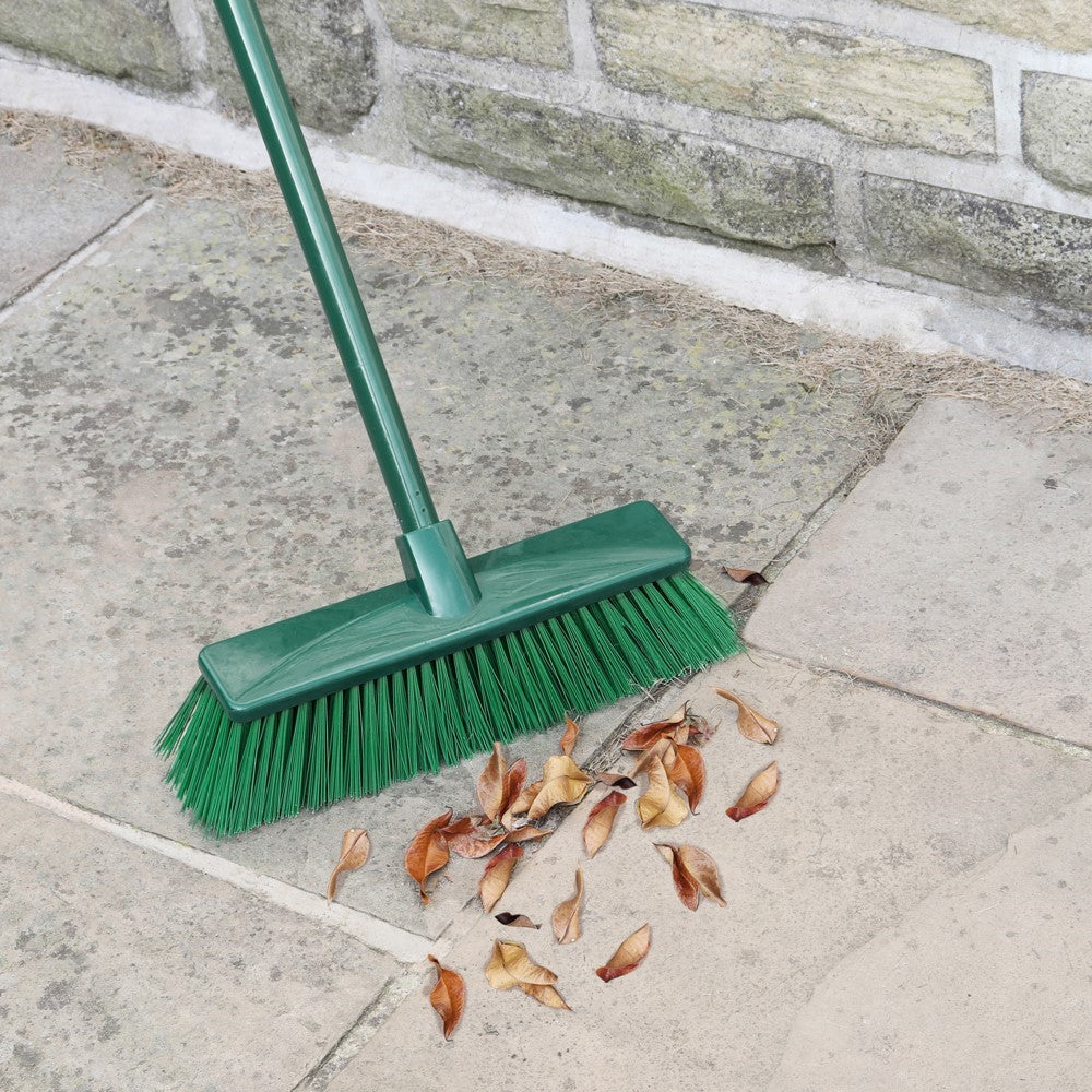 Green hard bristle broom with extendable handle sweeping dry leaves on stone patio near stone wall