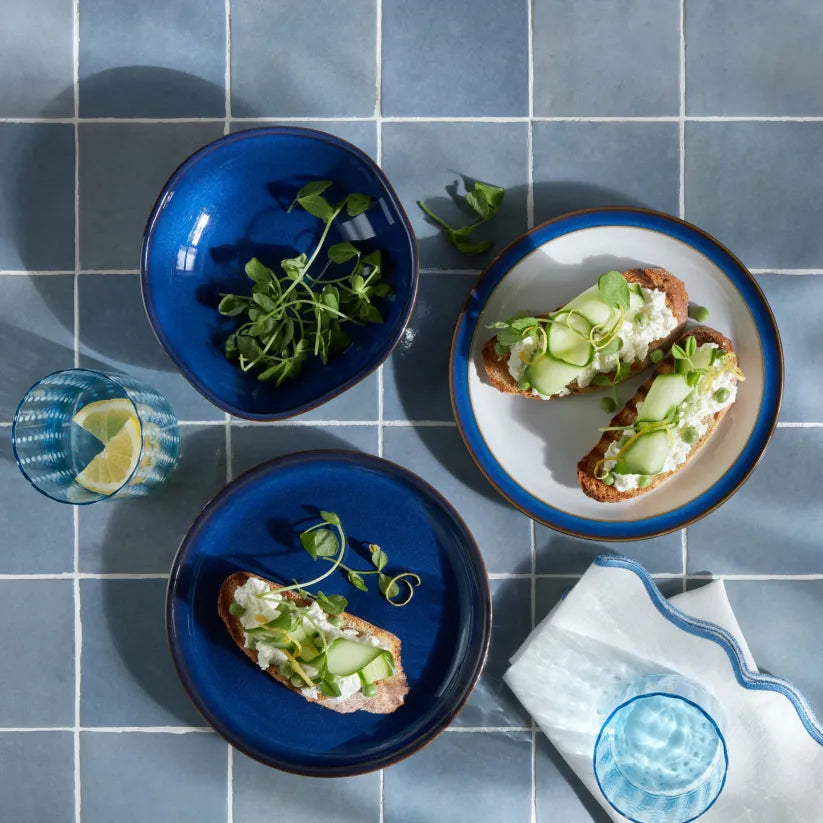 Denby Imperial Blue serving bowl with greens, alongside matching plates with cucumber-topped toast on blue tiled surface.