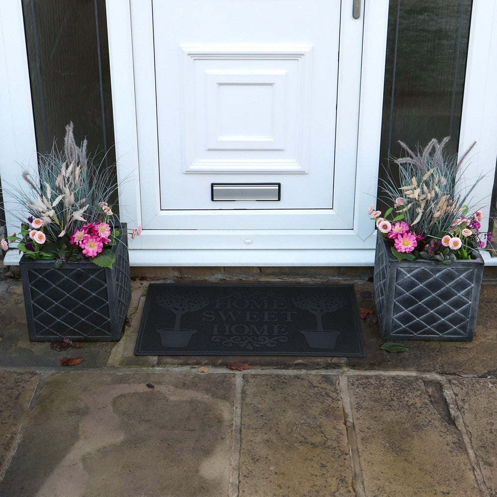 Black Latex Coir Home Sweet Home scraper mat with tree design placed outside a white door between two flower pots.