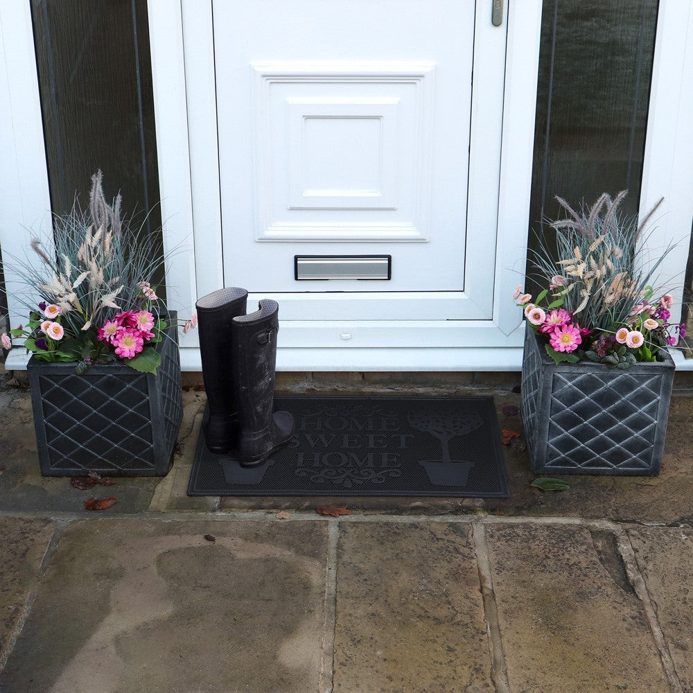 Black rubber scraper mat with "Home Sweet Home" text and plant pot designs placed outside a white door with boots nearby