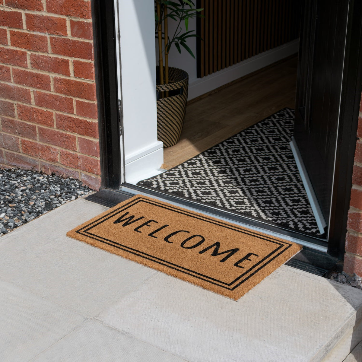 Latex-backed coir doormat with "WELCOME" text and border, placed outside an open black door.