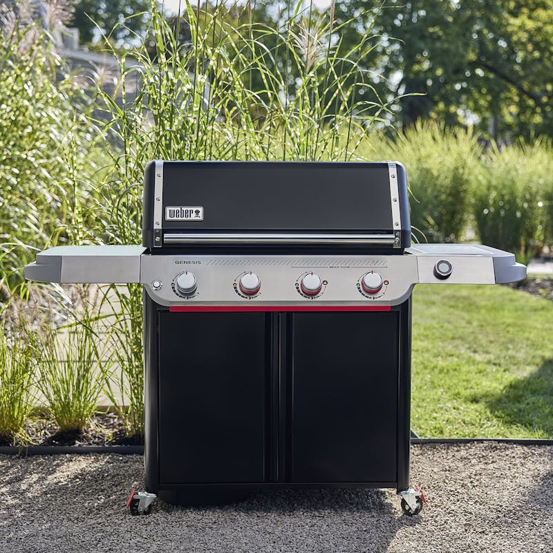 Genesis E-425W LP BLK gas barbecue with closed lid, four control knobs, and side shelves on a garden patio.
