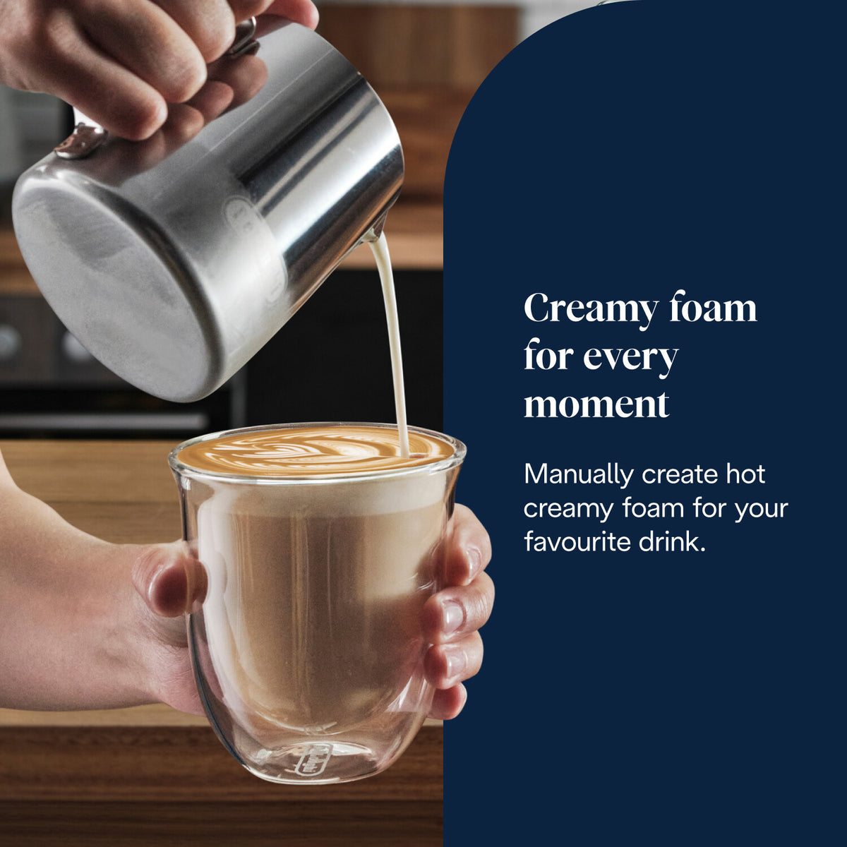Hand pouring creamy foam from a metal jug into a glass cup of coffee with latte art.