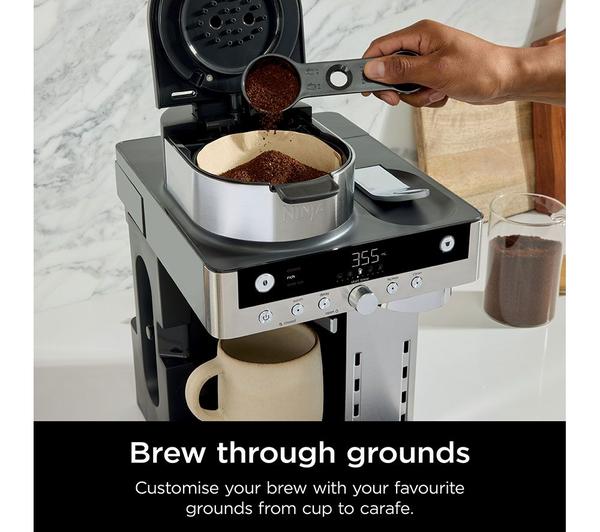 Ninja Prestige DualBrew coffee machine with open top, hand adding coffee grounds into filter basket above a beige mug.