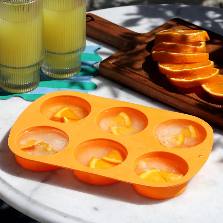 Tala orange silicone 6-hole ice mould with lemon slices frozen in ice on a marble table next to drinks and sliced oranges