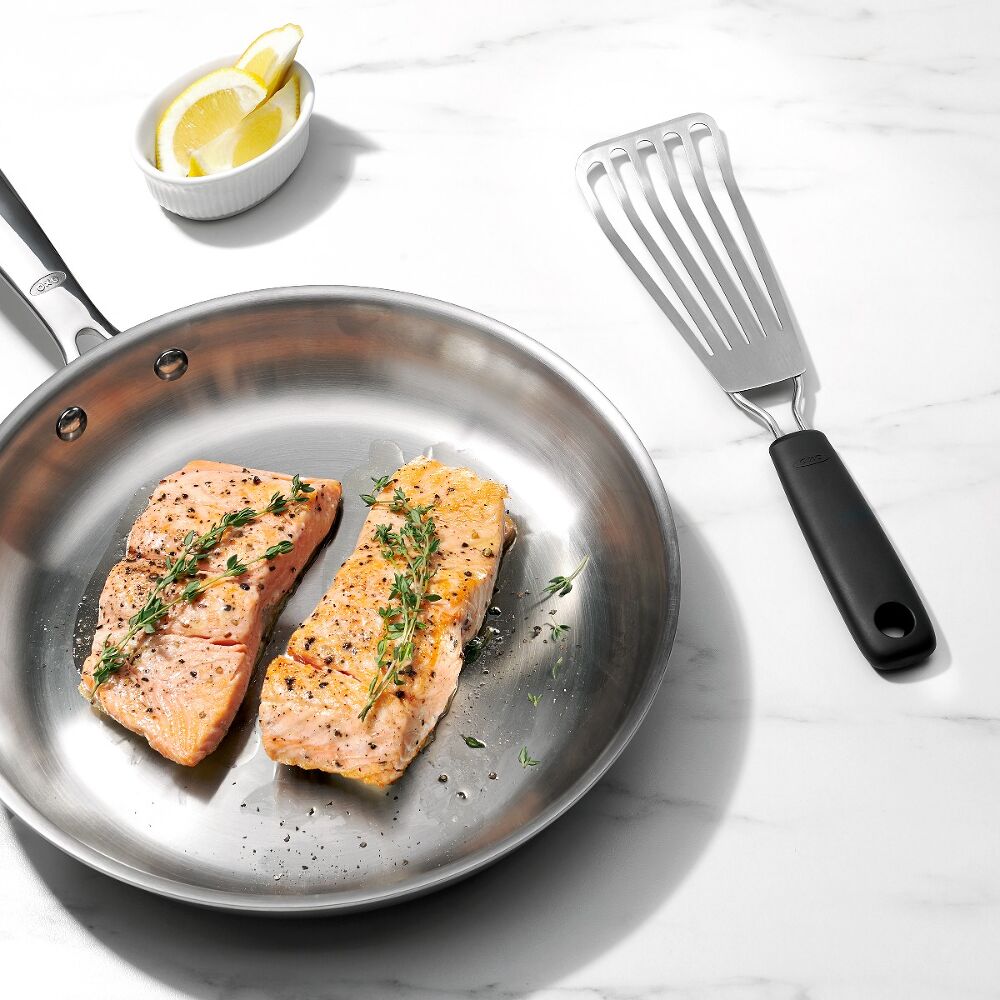 Oxo Little Fish Turner with black handle next to a stainless steel pan with two cooked salmon fillets and lemon wedges.