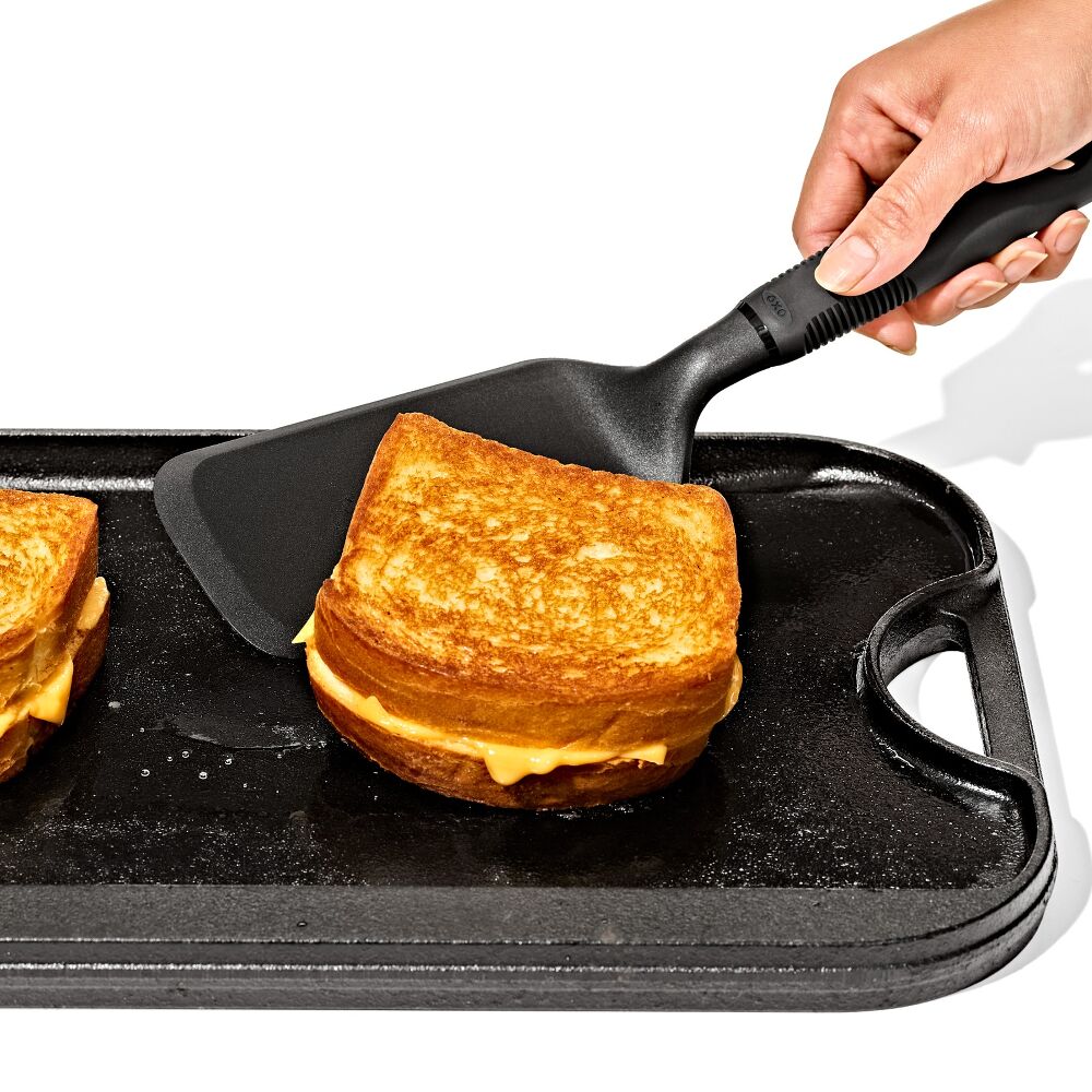 Hand holding Oxo Nylon Lasagne Turner lifting a grilled cheese sandwich on a griddle pan.