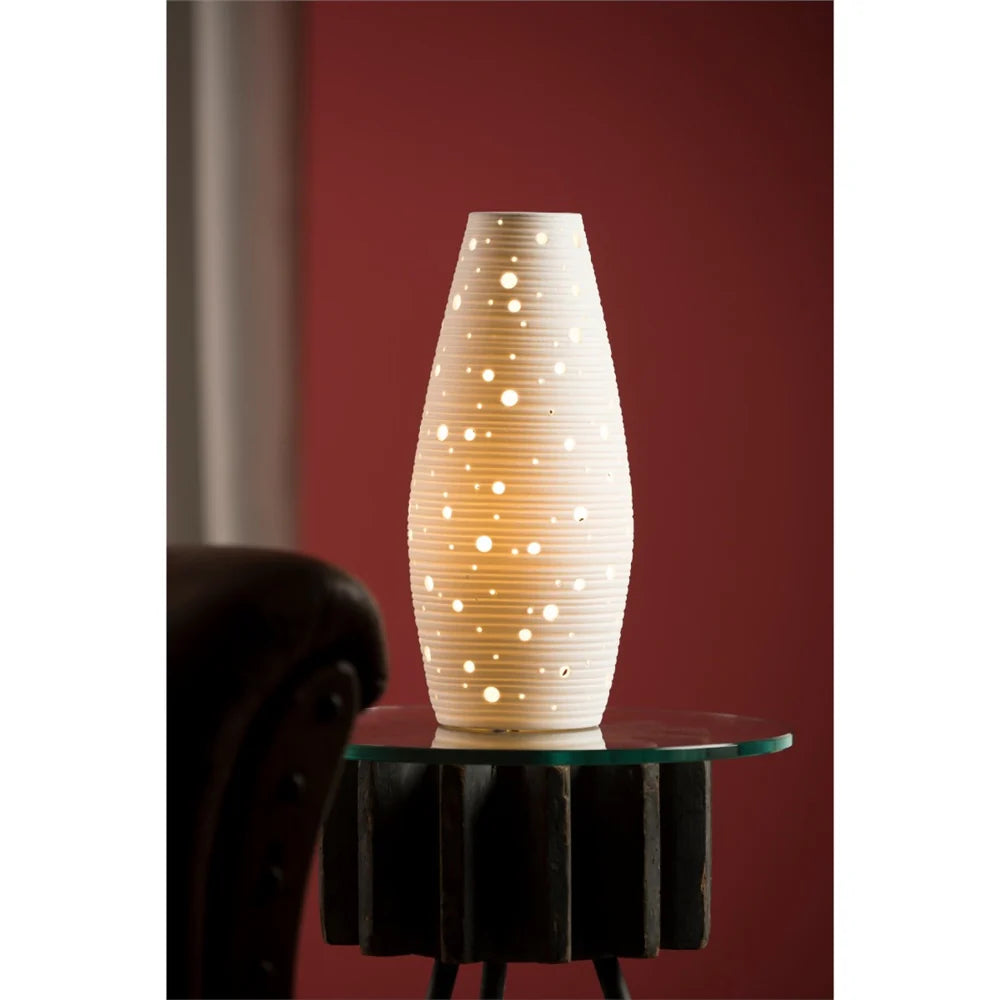 Glow Luminaire ceramic table lamp with perforated dots glowing warmly on a glass-topped wooden side table.