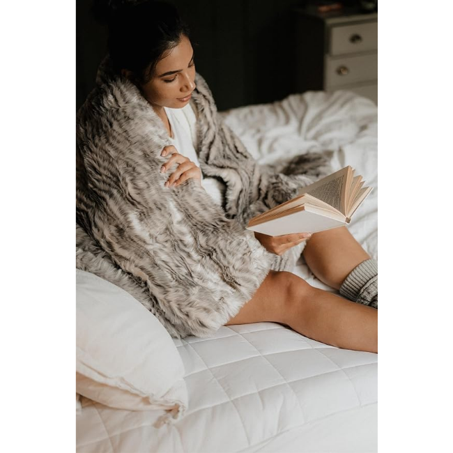 Double electric blanket with quilted white mattress cover on bed, woman wrapped in fur blanket reading a book