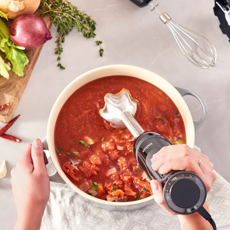 Braun Hand Blender MultiQuick 5 MQ5237BK blending tomato sauce in a pot with herbs and vegetables nearby.