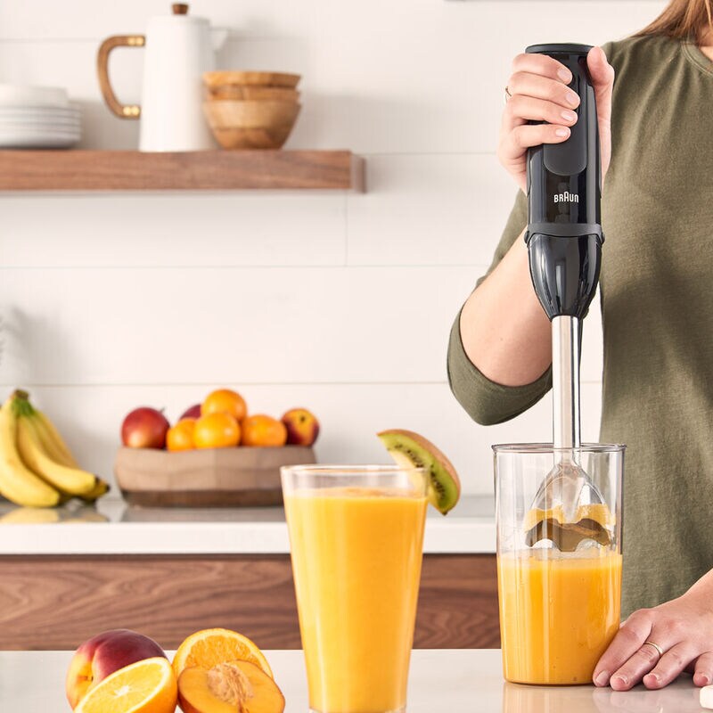 Person using Braun Hand Blender MultiQuick 5 MQ5237BK to blend orange juice in a clear container on a kitchen counter.