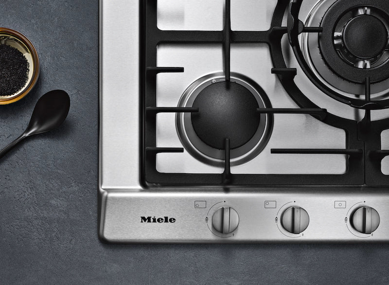 Close-up of Miele KM 2032 stainless steel gas hob corner with three control knobs and two burners visible.