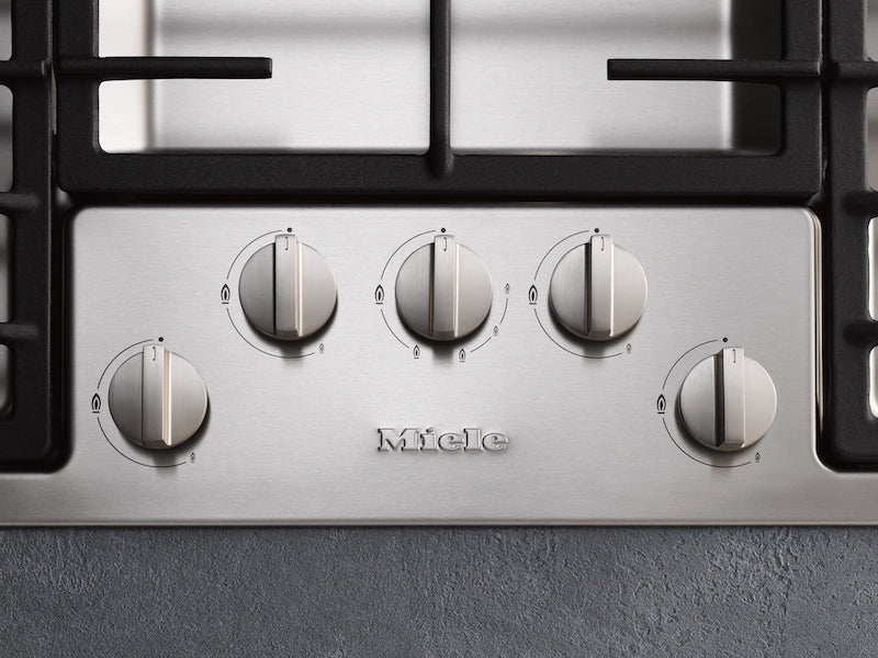 Close-up of Miele KM 2354-1 gas hob control panel with five stainless steel knobs and black grates above.