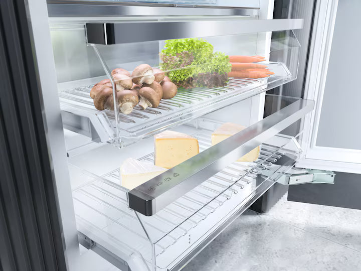 Open Miele K 2902 Vi fridge drawer showing mushrooms, lettuce, carrots, and cheese inside.