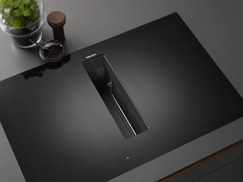 Miele KMDA 7676-1 FL black glass hob with integrated extractor vent in the center, shown on a dark countertop.