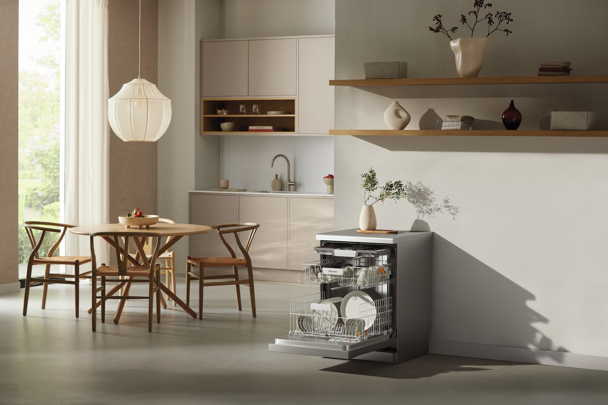 Open Miele G 5811 SC dishwasher with CleanSteel front in a modern kitchen setting next to a wooden dining table.