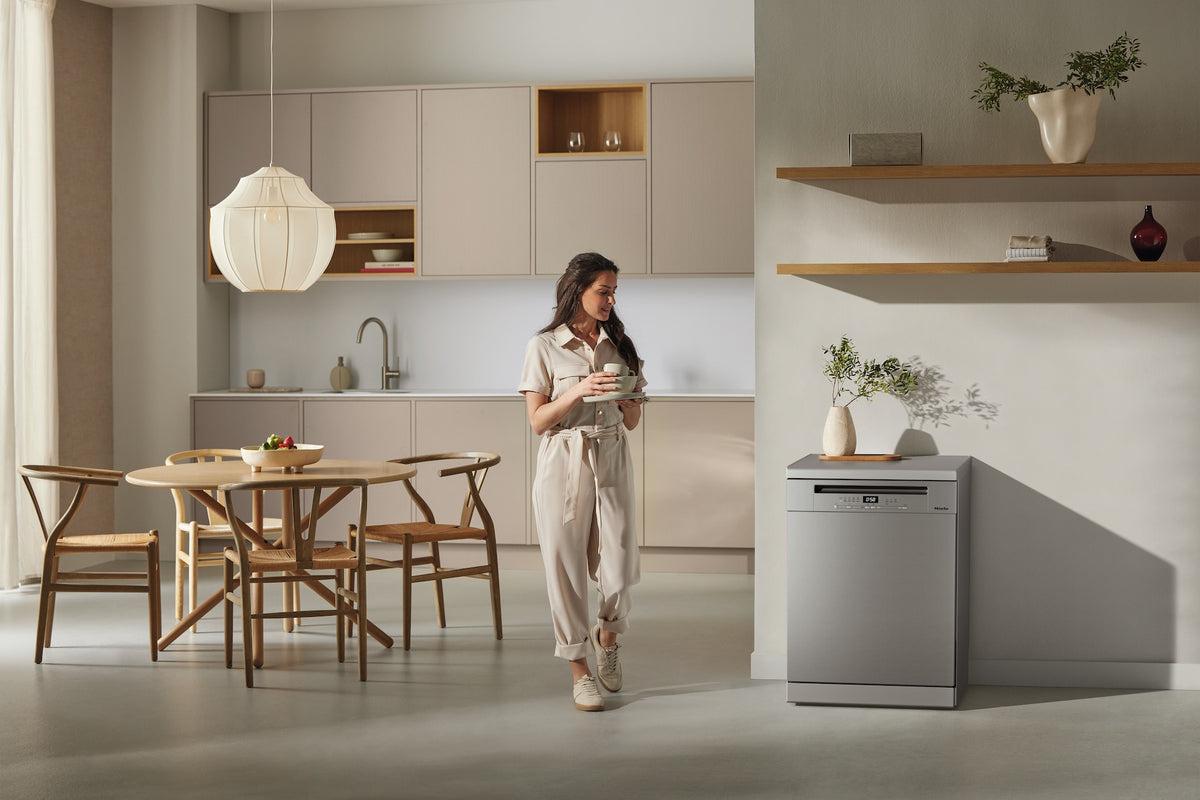 Miele G 5811 SC dishwasher with CleanSteel front in a modern kitchen setting next to wooden dining table and woman.