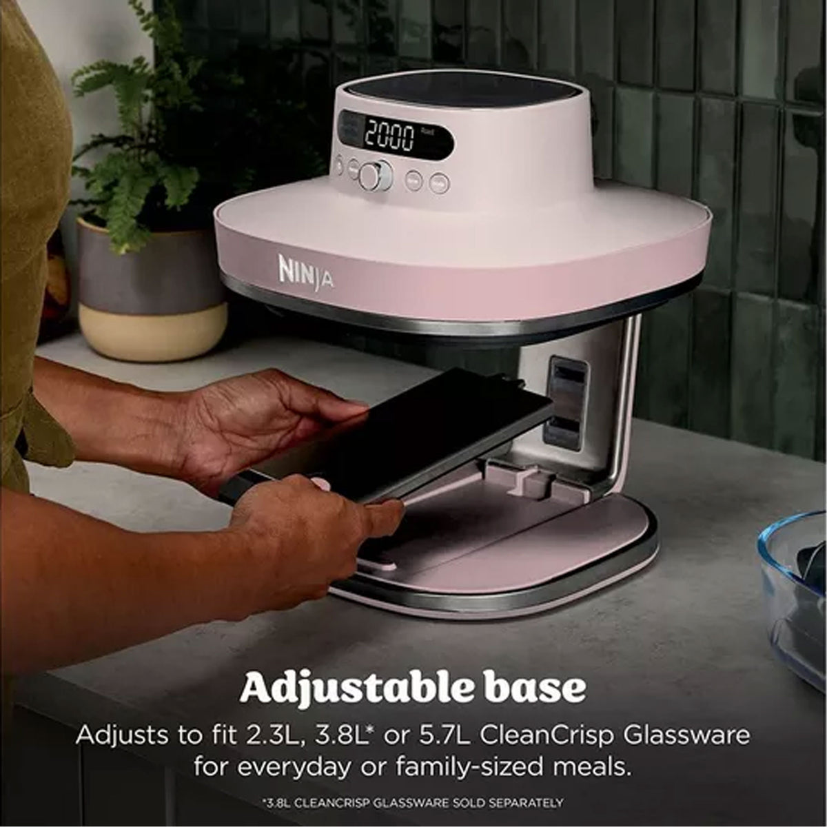 Hands adjusting the base of a rose quartz Ninja Crispi Pro 7-in-1 glass air fryer on a kitchen counter.