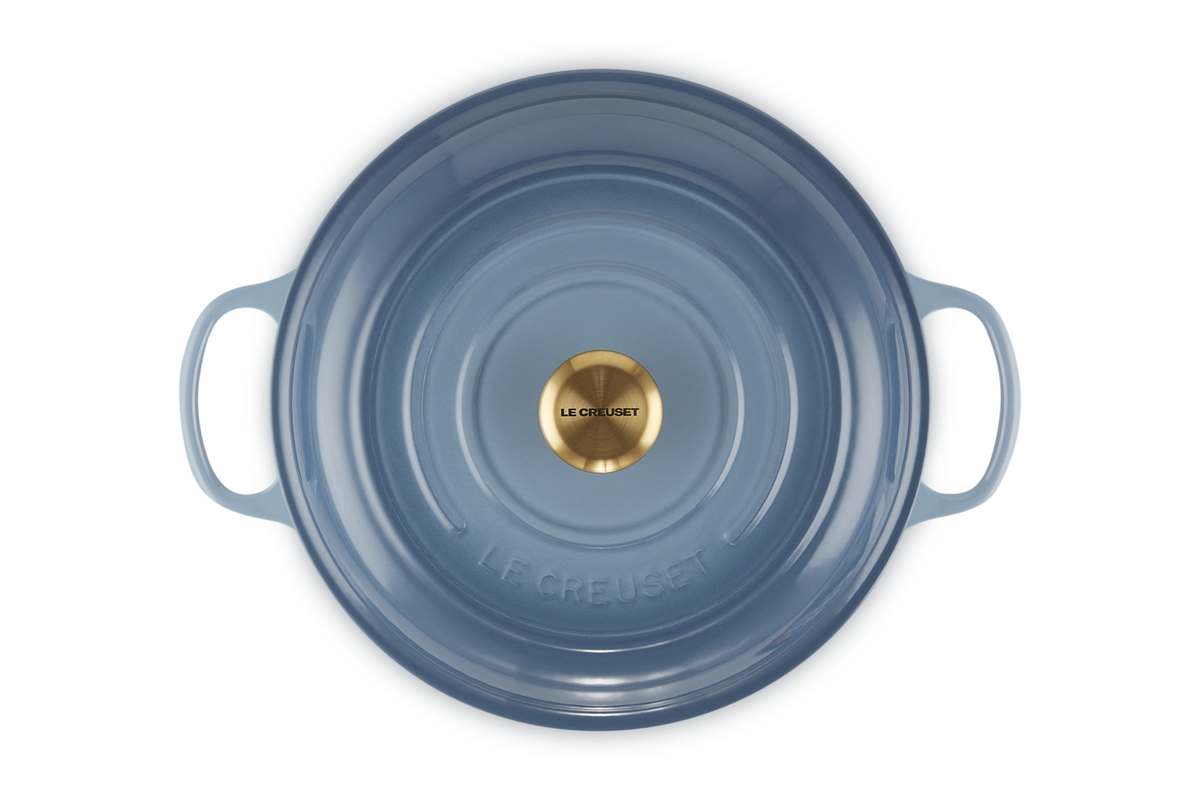 Le Creuset shallow cast iron casserole in chambray blue with gold knob, top view showing handles and lid details.