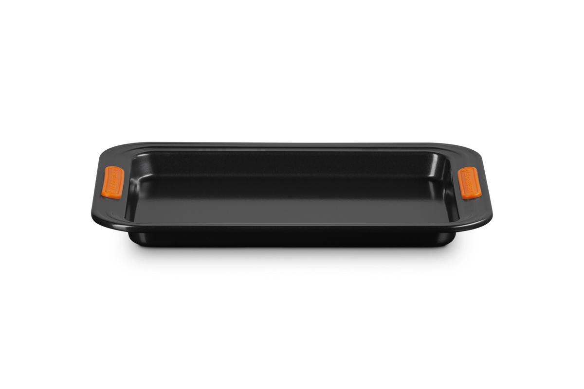 Le Creuset 32cm rectangular black baking sheet with orange silicone grip handles, front view.