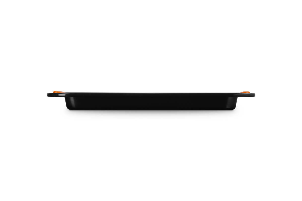 Side view of a black Le Creuset 32cm rectangular baking sheet with orange handles against a black background