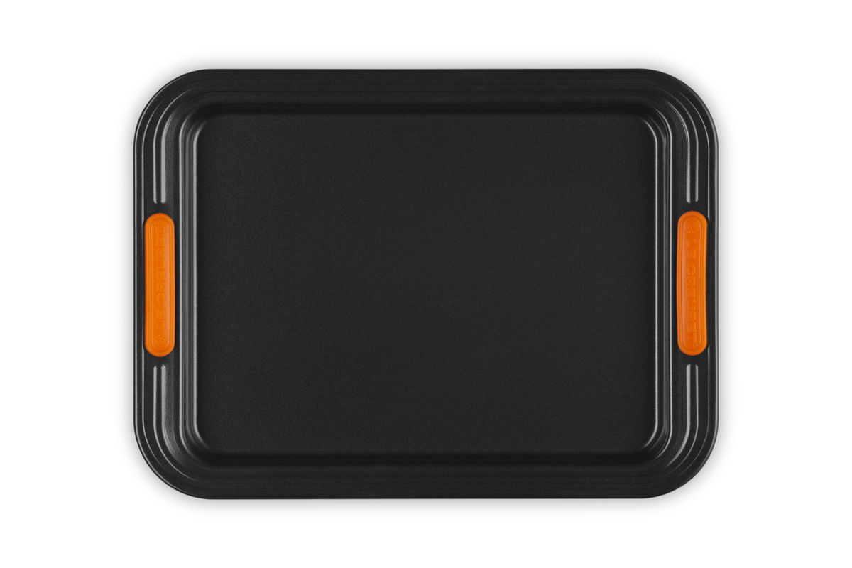 Le Creuset 32cm rectangular black baking sheet with orange silicone grip handles, top view.