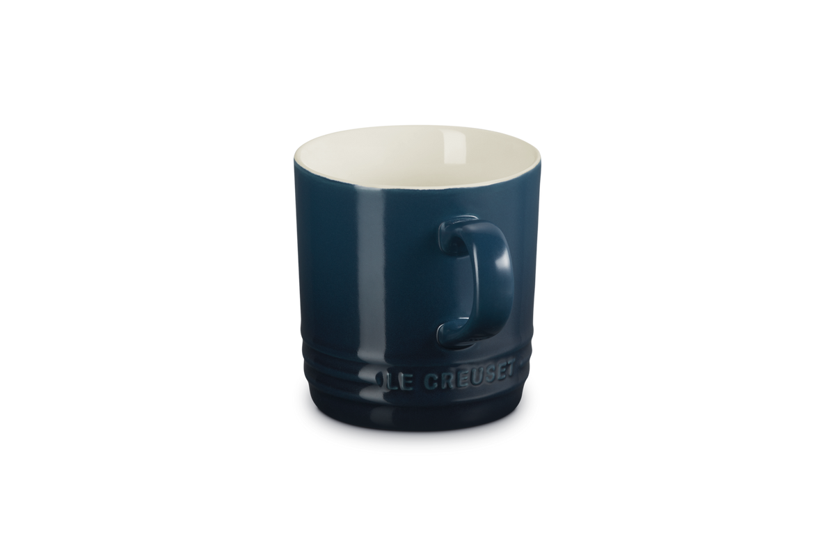 Le Creuset Cappuccino Mug in deep blue with white interior, handle visible, embossed logo near base.