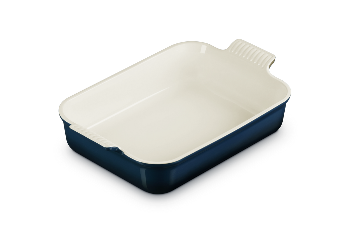 Le Creuset rectangular deep dish with cream interior and dark blue exterior, angled view showing handles.