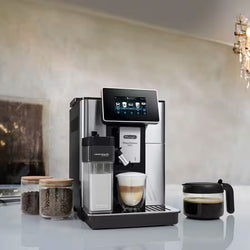 De'Longhi coffee machine with a glass of layered coffee and jars of coffee beans on a light surface.