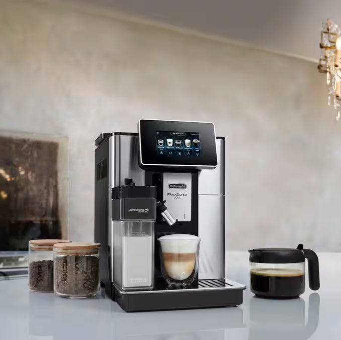 De'Longhi coffee machine with a glass of layered coffee and jars of coffee beans on a light surface.