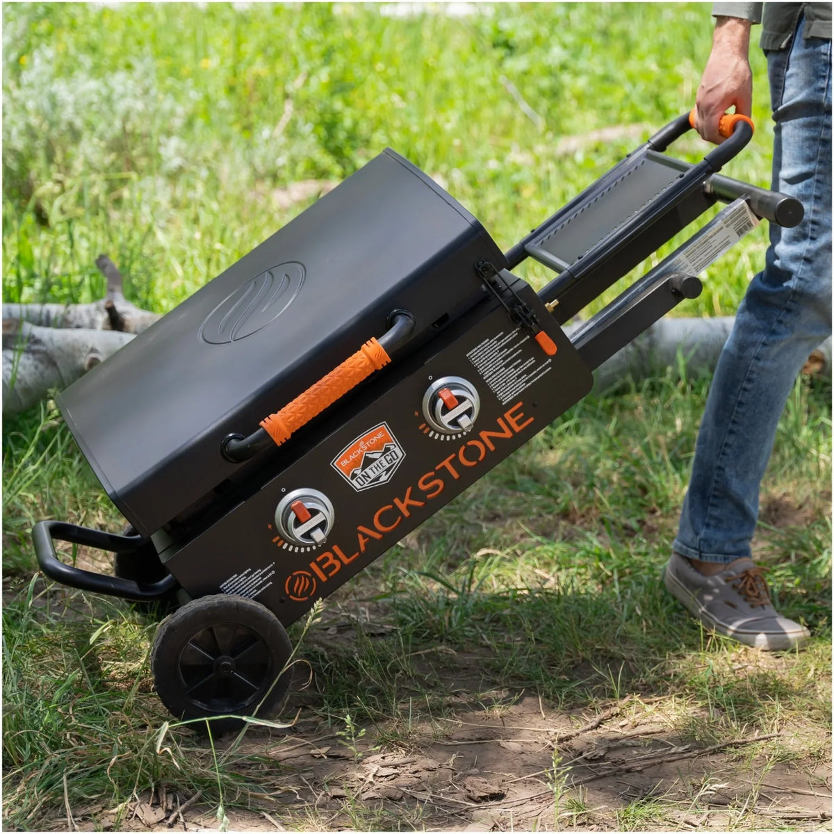 Person tilting Blackstone 22" On-The-Go Omnivore Flex-Fold Griddle with orange handles outdoors on grass