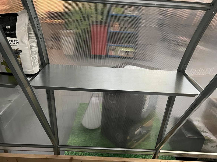 Galvanized steel greenhouse shelf installed inside a greenhouse with translucent walls and garden items visible outside.