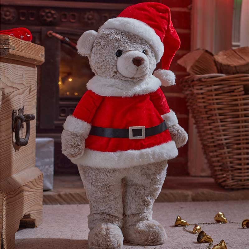 Teddy bear in a Santa outfit standing in a cozy room with a fireplace.