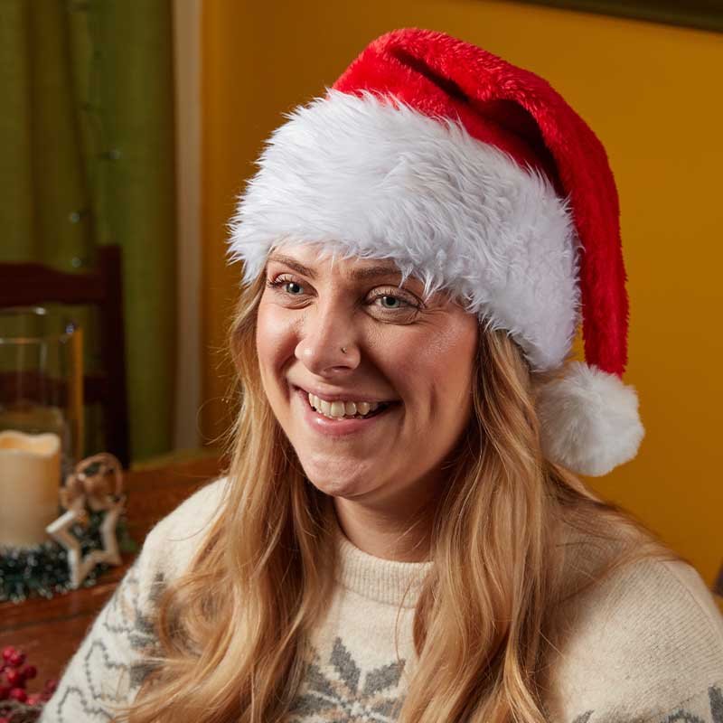 Woman smiling wearing a traditional red Santa hat with white trim and pom-pom indoors