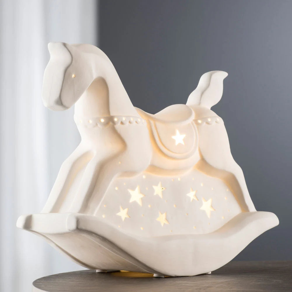 White ceramic rocking horse LED light with star-shaped cutouts glowing softly on a table.