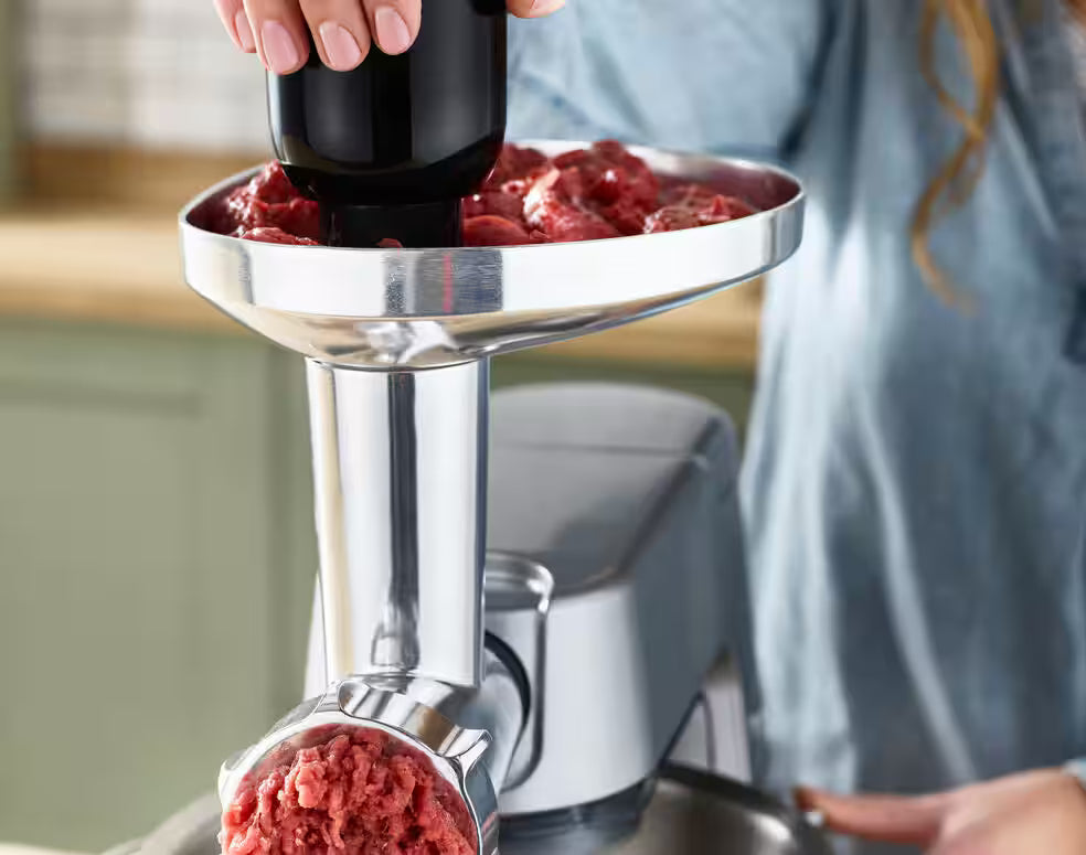 Kenwood Meat Grinder Attachment in use, grinding raw meat into a bowl below on a kitchen counter.