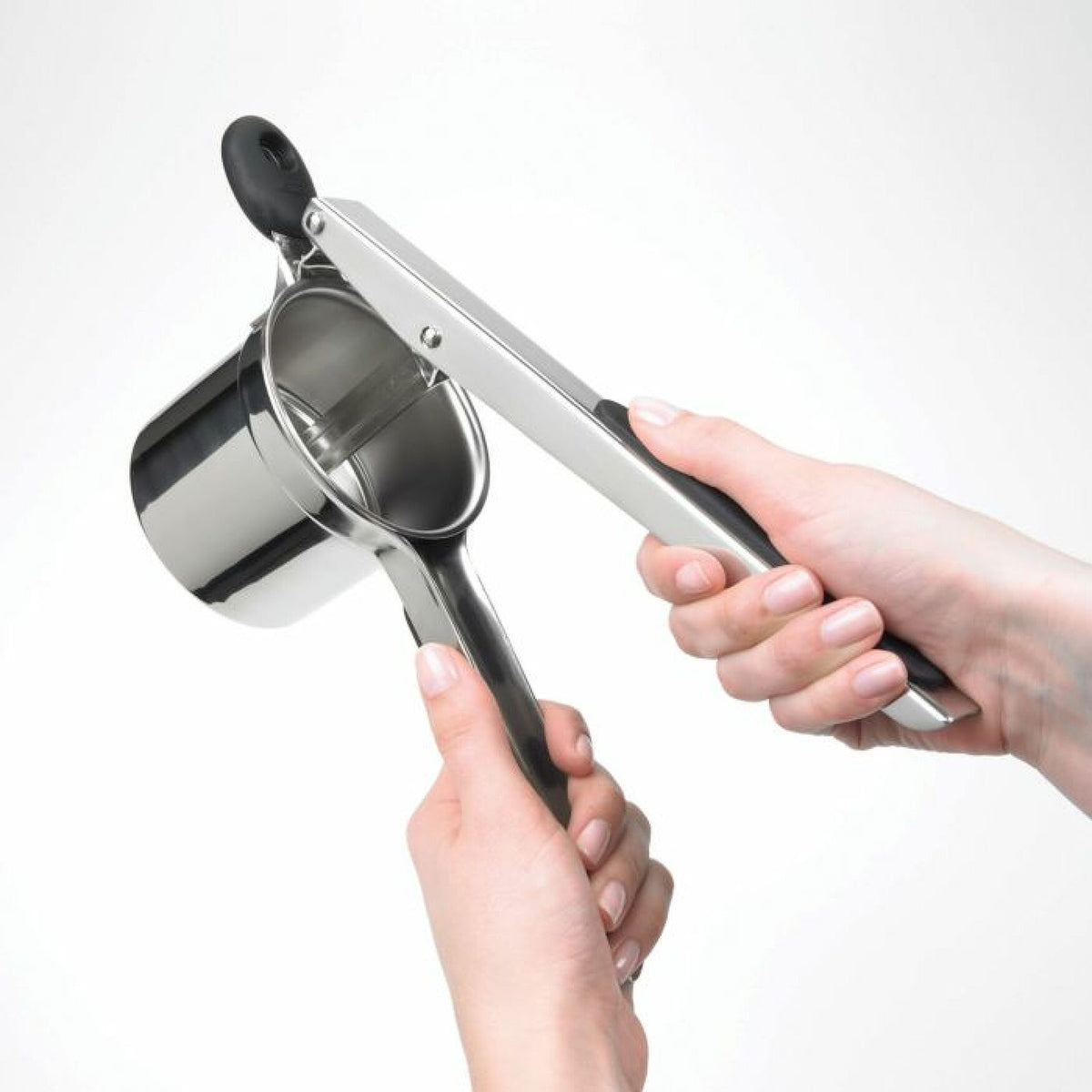 Hands holding an Oxo Potato Ricer with stainless steel basket and black handles against white background