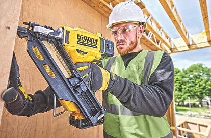 Man wearing safety gear using a DeWalt Twin Nailer Kit 18V on a wooden frame at a construction site.