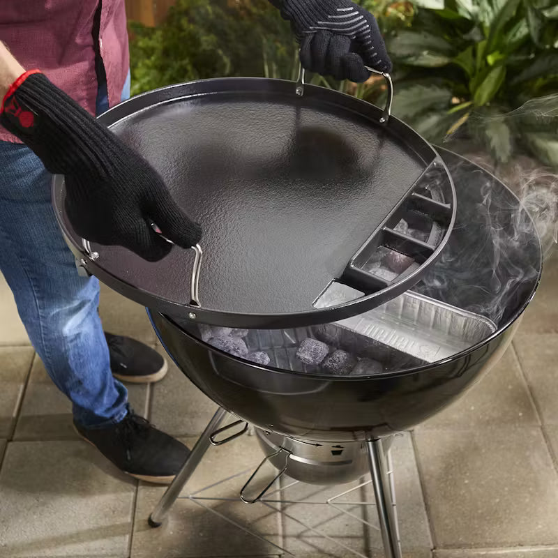 Person wearing gloves lifting a Weber full-size griddle off a charcoal 57cm kettle grill with hot coals inside.