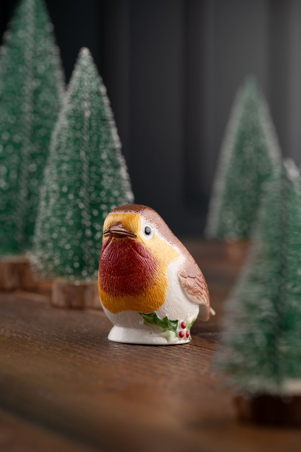 Close-up of a ceramic robin ornament with holly detail, surrounded by blurred miniature Christmas trees.
