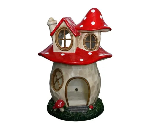 A decorative miniature fairy house shaped like a mushroom, featuring a red and white spotted cap-shaped roof, a beige stone-textured body, arched door and windows with gold trim, and a small green grassy base with tiny red mushrooms.