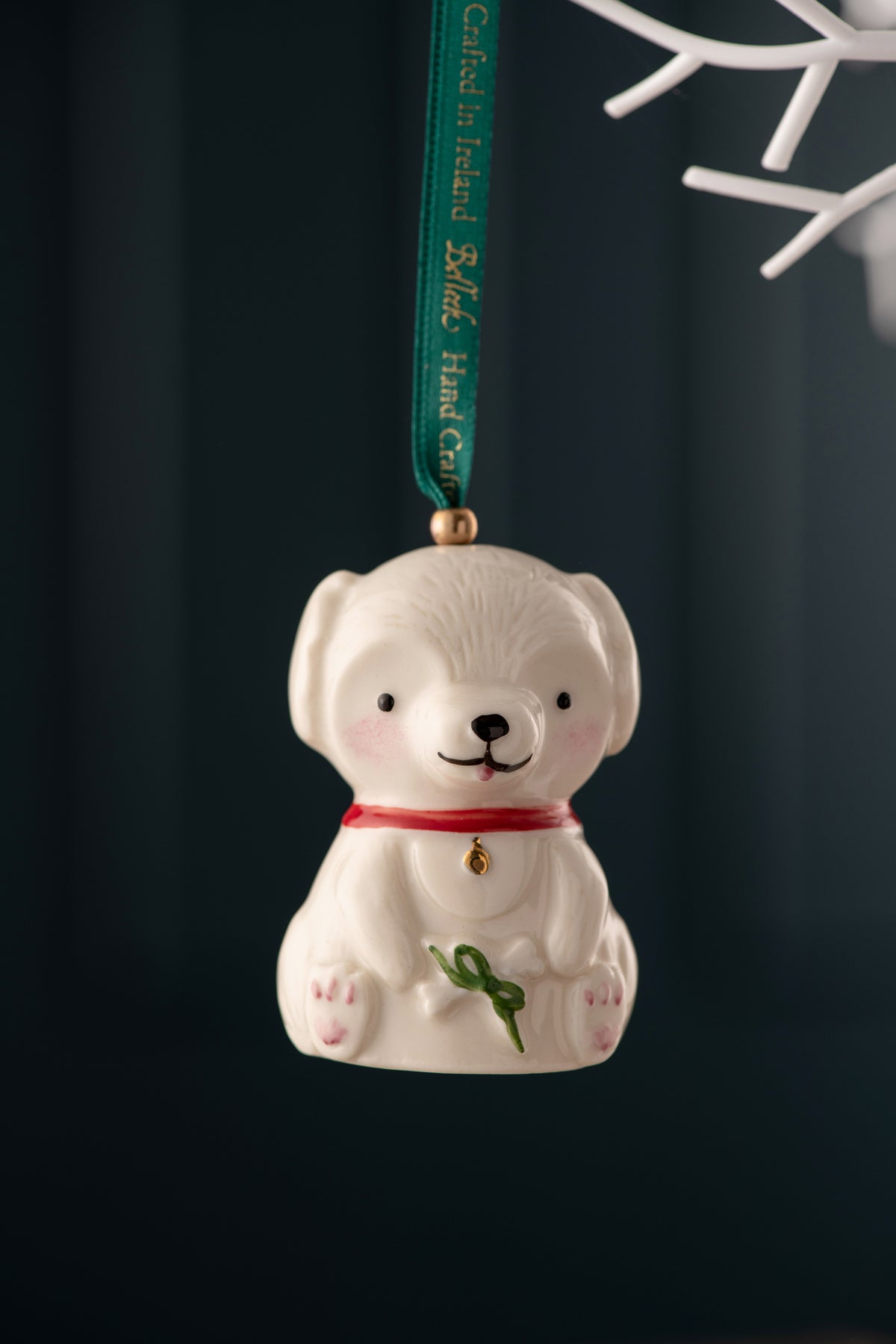 White ceramic dog ornament with red collar and green bow, hanging from a green ribbon against a dark background
