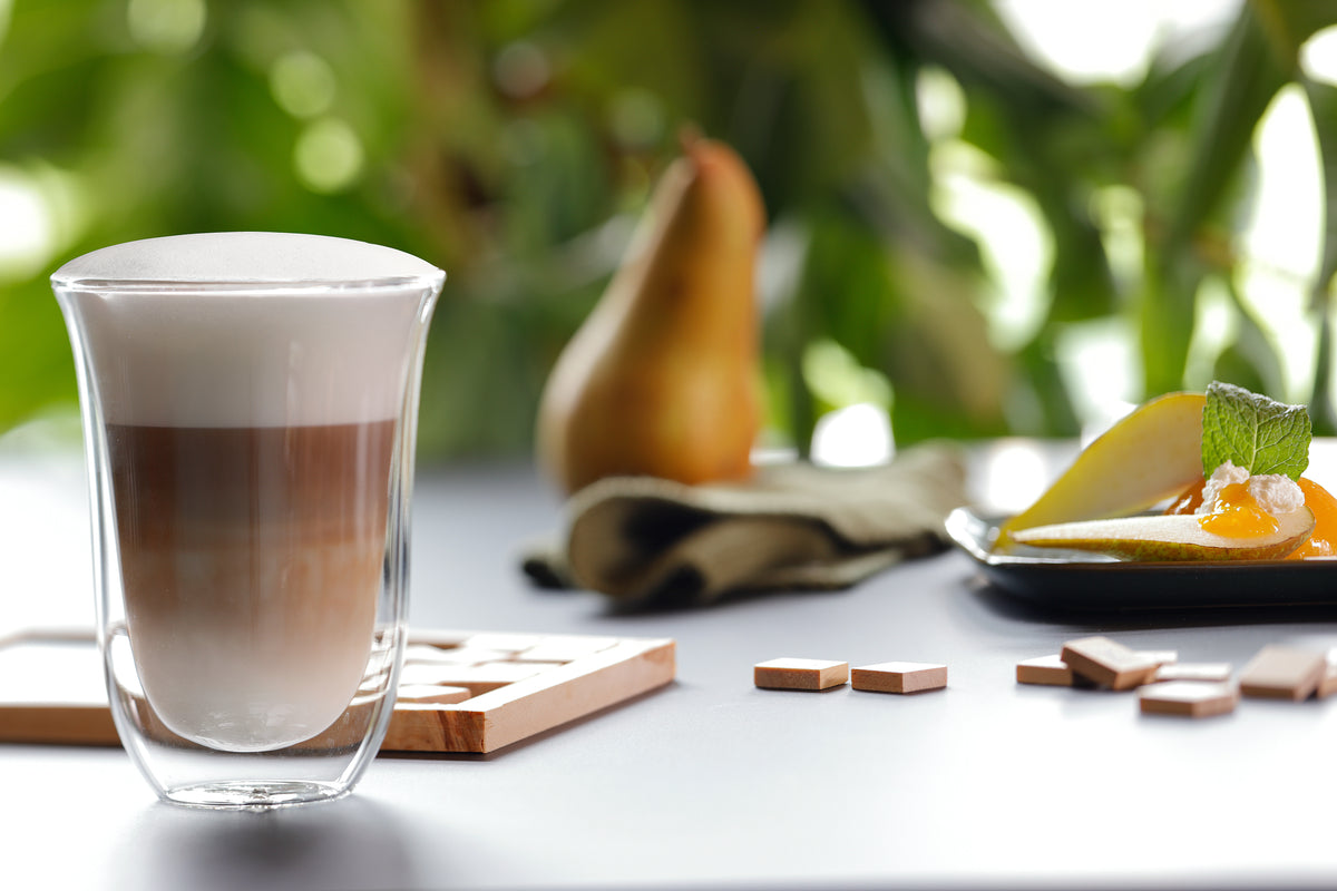 DeLonghi double wall glass filled with layered latte macchiato, blurred pear and dessert in background