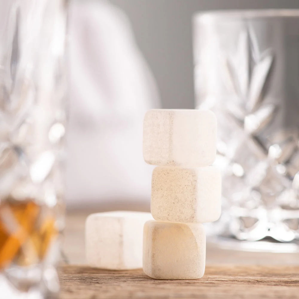 Four white jade cooling stones stacked and resting on a wooden surface with blurred glassware in the background.