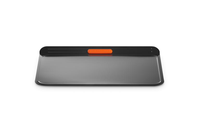 Le Creuset insulated cookie sheet with a black handle and orange grip, shown flat on a black background.