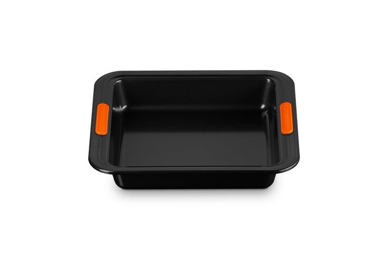 Le Creuset square black non-stick cake tin with orange heat-resistant silicone handles, top view.