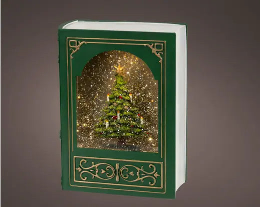 A green hardcover book-shaped snow globe with gold decorative borders and a miniature Christmas tree inside, shown against a dark gray gradient background.