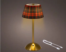 A table lamp with a gold-finished metal base and stem, featuring a pleated fabric shade in a red, black, and gold tartan plaid pattern. The lamp is shown illuminated against a dark gray gradient background, with a white USB charging cable displayed to the right.