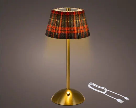 A table lamp with a gold-finished metal base and stem, featuring a pleated fabric shade in a red, black, and gold tartan plaid pattern. The lamp is shown illuminated against a dark gray gradient background, with a white USB charging cable displayed to the right.
