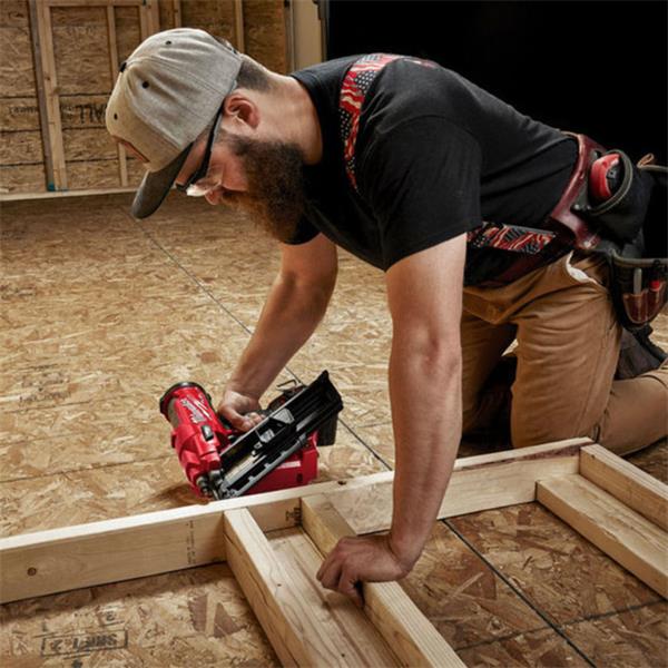 Man kneeling on plywood floor using Milwaukee M18 FUEL cordless framing nailer on wooden frame.